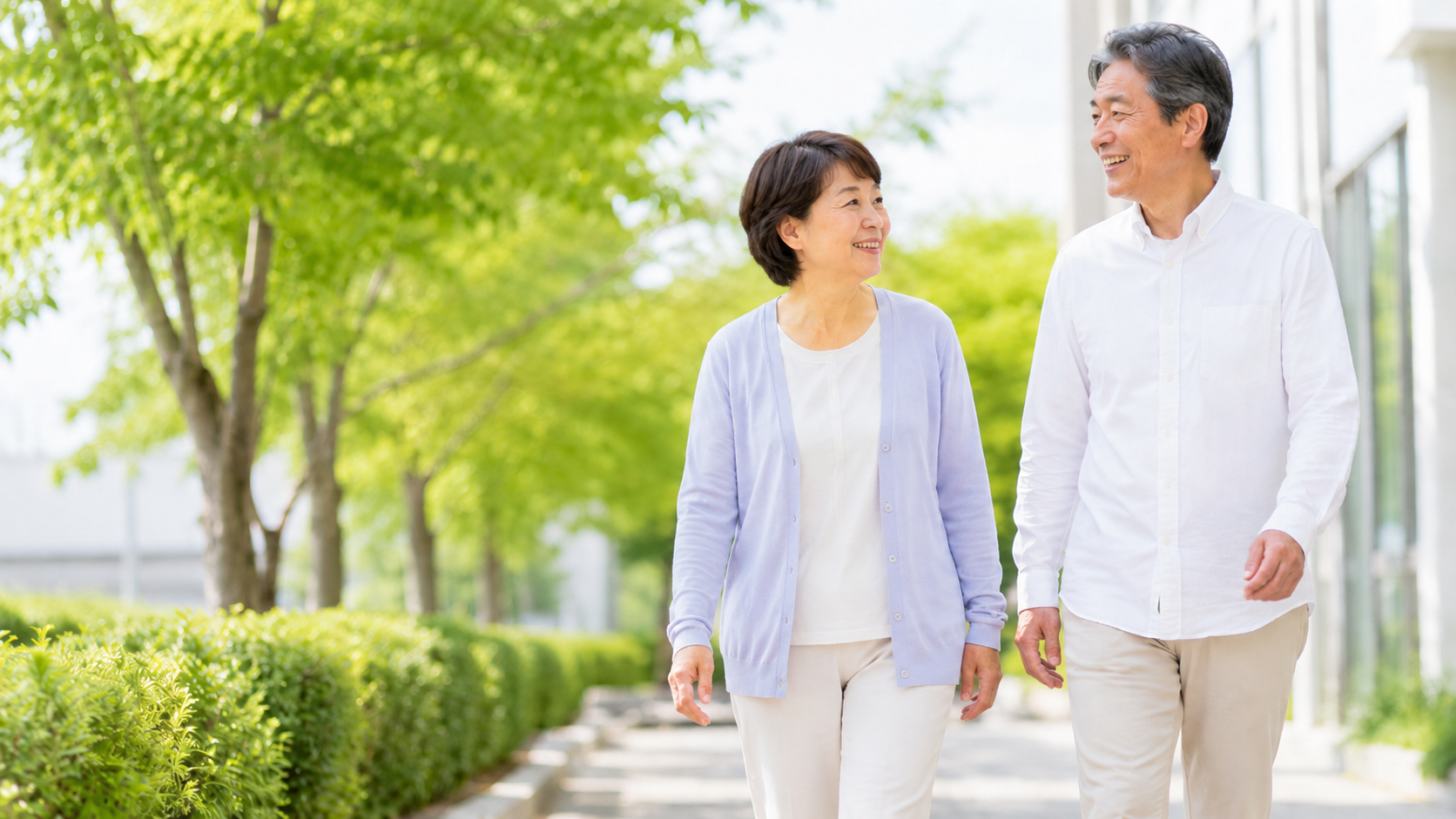 A smiling couple strolling down a sunlit path beside green trees and shrubs.